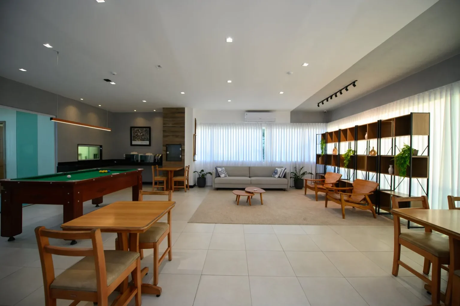 Salão de convivência iluminado com mesa de sinuca verde, sala de estar e estantes