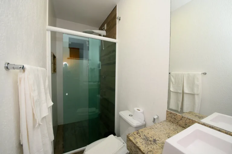 Banheiro com box de vidro, bancada de granito, cuba branca, espelho e toalhas do hotel