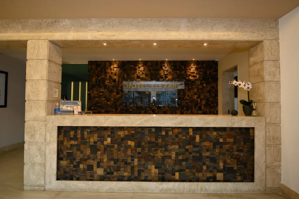 Balcão da recepção do Hotel Elevare com painel de madeira em mosaico, letreiro dourado e orquídea