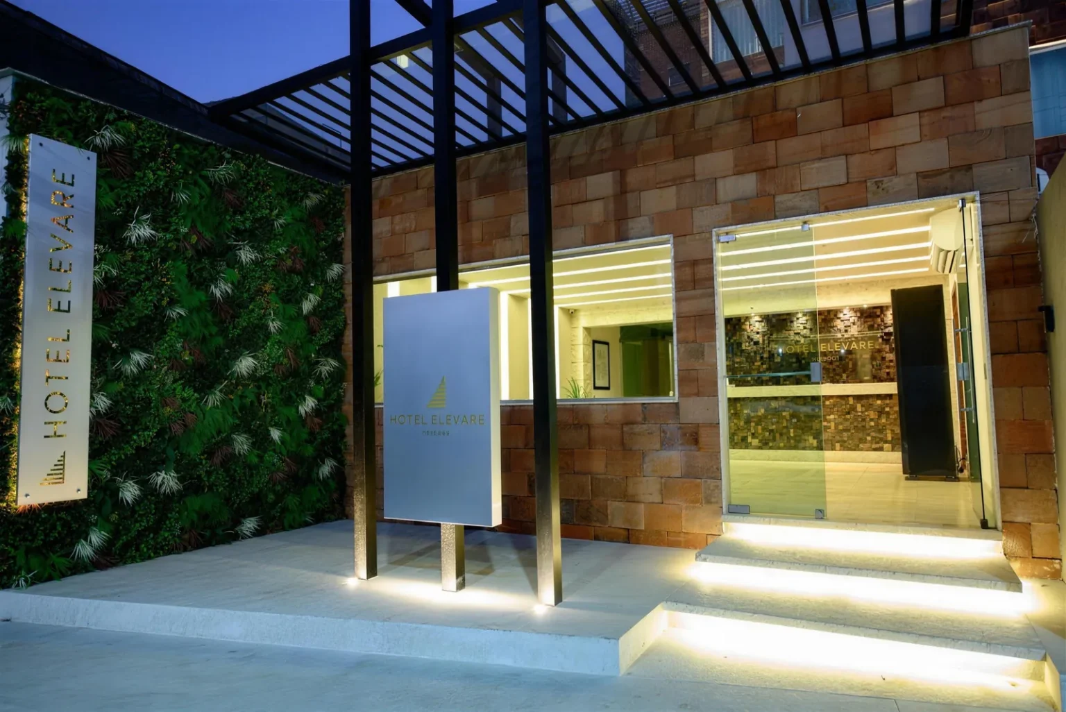 Entrada do Hotel Elevare Niterói ao entardecer, com parede de pedra, jardim vertical e totem iluminado