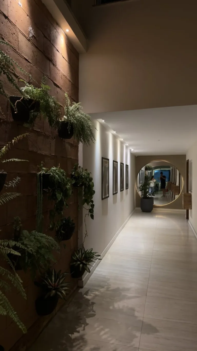 Corredor com parede de tijolinho, jardim vertical de samambaias e espelho redondo
