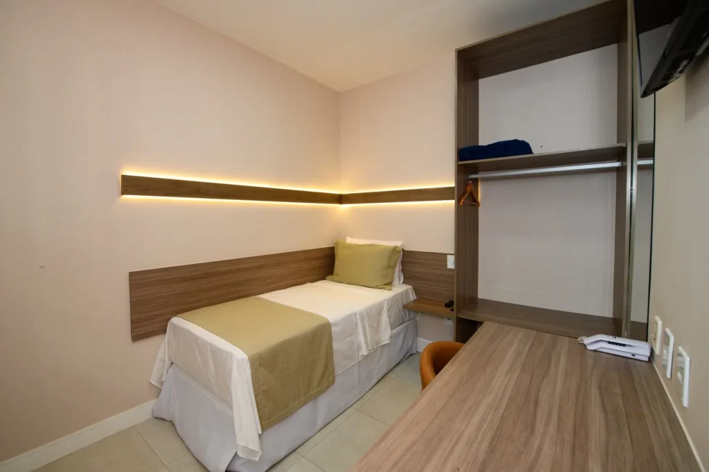 Quarto Standard Solteiro com cama arrumada, cabeceira com luz indireta, armário aberto e bancada de trabalho