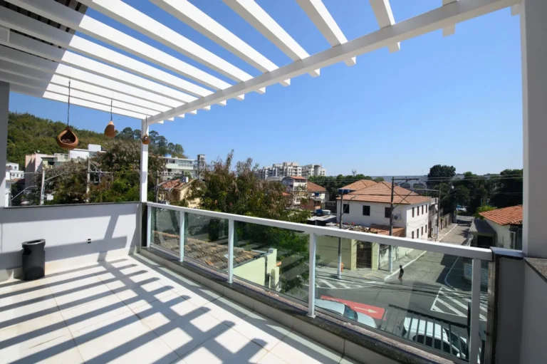 Terraço com pergolado branco e guarda-corpo de vidro, com vista do bairro em dia de sol